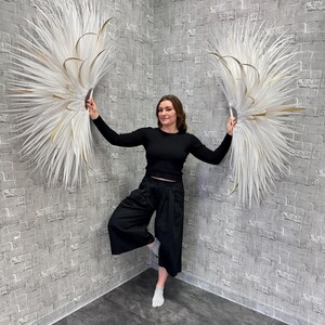 May include: Two large, white, feathery wall decorations with gold accents are held up by a person. The decorations are in a fan shape and are against a gray brick wall. The person is wearing a black long-sleeve shirt and black pants.