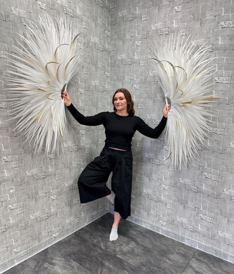 May include: Two large, white, feathery wall decorations with gold accents are held up by a person. The decorations are in a fan shape and are against a gray brick wall. The person is wearing a black long-sleeve shirt and black pants.