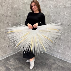 May include: A white tutu with gold accents, held by a person wearing a black dress. The tutu has a circular shape with long, spiky extensions radiating outwards. The gold accents are visible along the edges of the tutu.