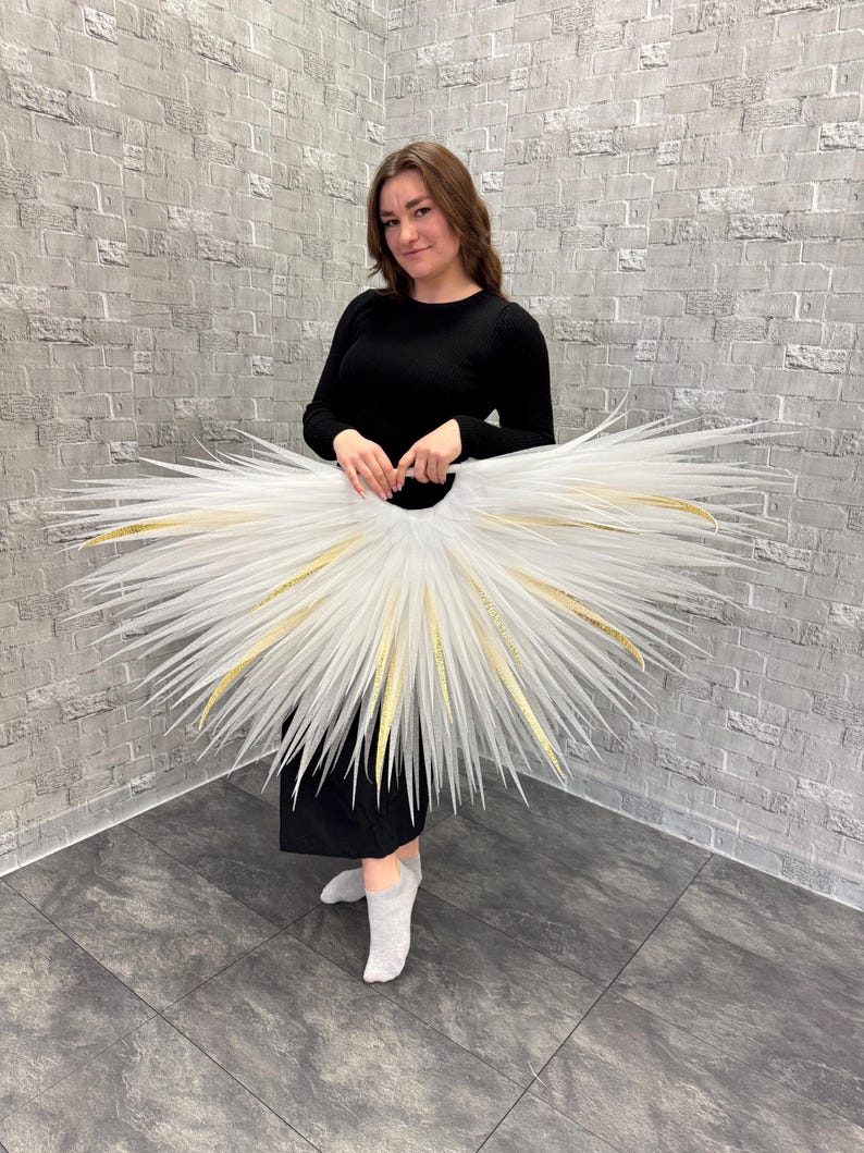May include: A white tutu with gold accents, held by a person wearing a black dress. The tutu has a circular shape with long, spiky extensions radiating outwards. The gold accents are visible along the edges of the tutu.