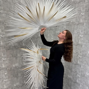 May include: Two large, white feather fans with gold accents are held up against a gray brick wall. The fans have a radial design with long, pointed feathers. The gold accents are in the form of long, thin, gold-colored feathers. The fans are held by a person wearing a black dress.