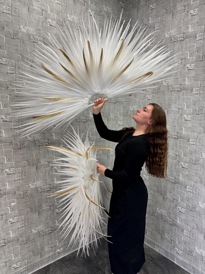 May include: Two large, white feather fans with gold accents are held up against a gray brick wall. The fans have a radial design with long, pointed feathers. The gold accents are in the form of long, thin, gold-colored feathers. The fans are held by a person wearing a black dress.