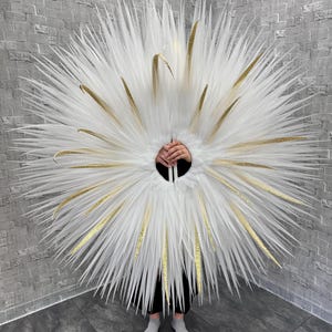 May include: A large, circular costume piece with white, feathery plumes and gold accents. The design features a central opening and is worn by a person. The background is a gray brick wall.