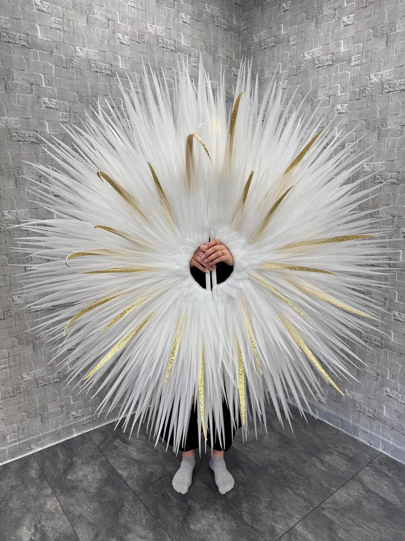 May include: A large, circular costume piece with white, feathery plumes and gold accents. The design features a central opening and is worn by a person. The background is a gray brick wall.