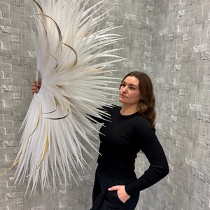 May include: A large, decorative white and gold floral arrangement. The arrangement features long, feathery white elements with gold accents. The piece is held against a gray brick wall, creating a striking contrast. The overall aesthetic is modern and elegant.