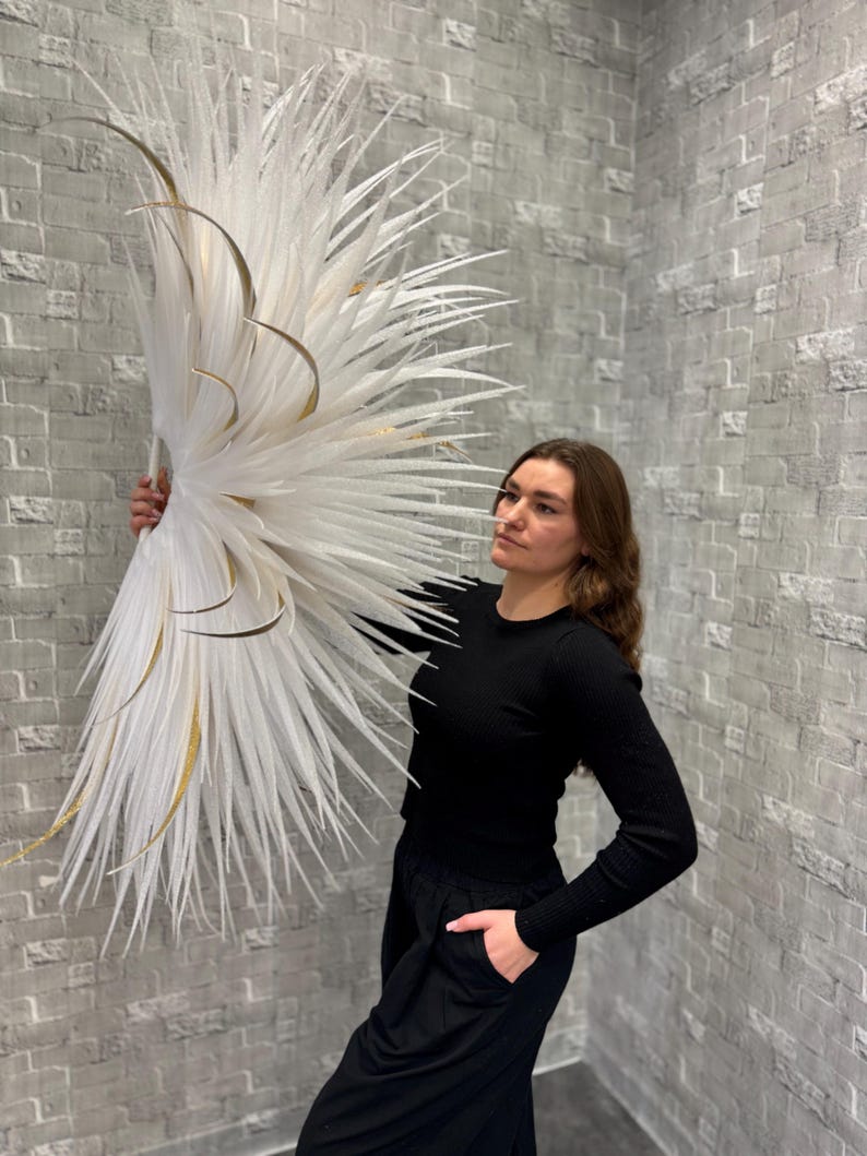 May include: A large, decorative white and gold floral arrangement. The arrangement features long, feathery white elements with gold accents. The piece is held against a gray brick wall, creating a striking contrast. The overall aesthetic is modern and elegant.