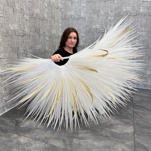 May include: A large, white, feathery decorative piece with gold accents. The piece is held by a person in a black shirt. The feathers radiate outward, creating a fan-like shape. The gold accents are curved and add a touch of elegance.