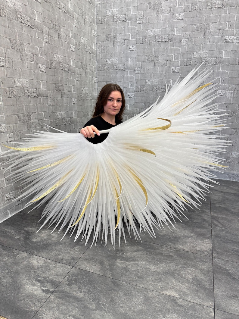 May include: A large, white, feathery decorative piece with gold accents. The piece is held by a person in a black shirt. The feathers radiate outward, creating a fan-like shape. The gold accents are curved and add a touch of elegance.