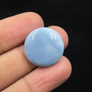 May include: A round, polished gemstone in a soft, light blue hue. The stone has a smooth, glossy surface with subtle internal patterns. The gemstone is held in a hand, with the background out of focus.