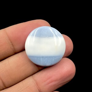 May include: A round, polished gemstone with a layered appearance. The stone features bands of light blue and white, creating a soft, marbled effect. The gemstone is held in a hand against a black background.