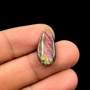 May include: A teardrop-shaped labradorite gemstone with a polished surface. The stone displays a vibrant play of color, with hues of purple, pink, and gold. The gemstone is held in a hand against a black background.