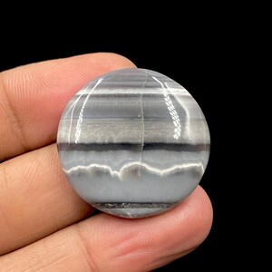 May include: A round, polished gemstone with a banded pattern of gray, white, and black. The stone is held in a hand against a black background. The bands create a layered effect, with a wavy white line at the bottom.