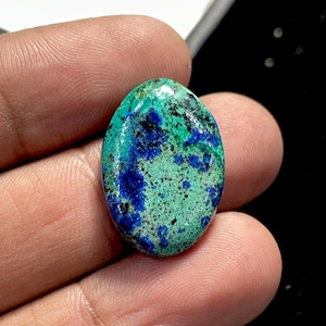 May include: Oval-shaped gemstone with a vibrant mix of colors. The stone features a base of teal and green, speckled with dark spots, and accented with deep blue patches. The polished surface reflects light, highlighting the natural patterns and textures of the stone.