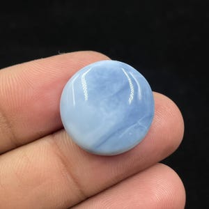 May include: A round, polished gemstone with a light blue and white color gradient. The stone has a smooth, glossy surface and is held in a person's hand against a dark background. The gemstone is a cabochon, likely for jewelry making.