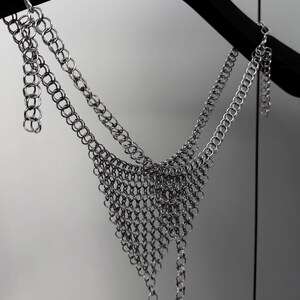 May include: A silver chainmail top hangs from a black hanger. The top features a triangular design with multiple rows of interconnected metal rings. The chainmail is made of small, shiny, silver-colored rings.