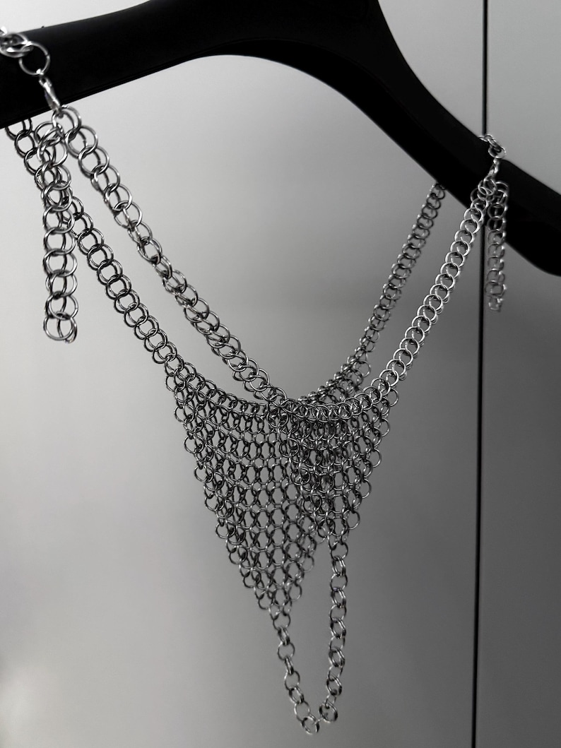 May include: A silver chainmail top hangs from a black hanger. The top features a triangular design with multiple rows of interconnected metal rings. The chainmail is made of small, shiny, silver-colored rings.