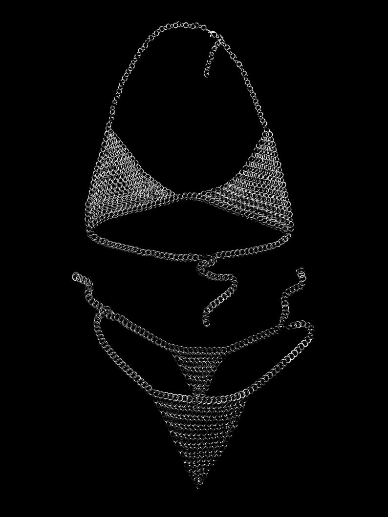 May include: A silver chainmail bikini set against a black background. The set includes a halter top and a thong-style bottom, both constructed from small, interlocking metal rings. The chains are reflective, creating a shimmering effect.
