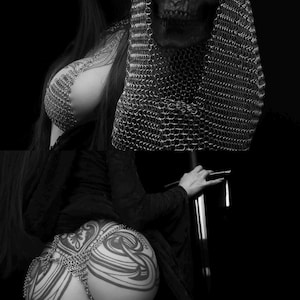 May include: Black and white image featuring a skull wearing a chainmail hood. The image also shows a person with tattoos and chainmail body jewelry. The person is holding a sword. The overall theme is gothic and dark.