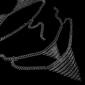 May include: A silver chainmail bikini top and chainmail body jewelry against a black background. The chainmail is made of small, interlocking metal rings, creating a shimmering effect. The design is intricate and detailed.
