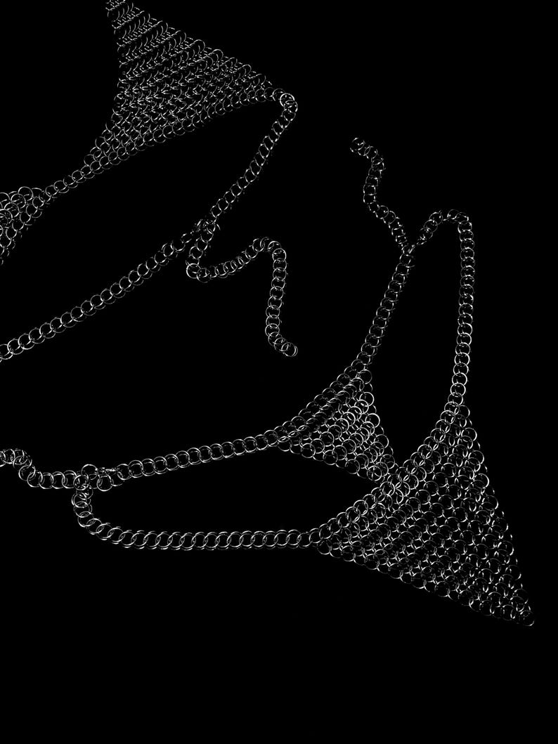 May include: A silver chainmail bikini top and chainmail body jewelry against a black background. The chainmail is made of small, interlocking metal rings, creating a shimmering effect. The design is intricate and detailed.