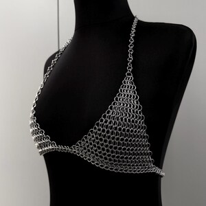 May include: A silver chainmail bikini top displayed on a black mannequin. The top features a halter neck design and triangular cups, constructed entirely of small, interlocking metal rings. The metallic garment is shown against a neutral background.