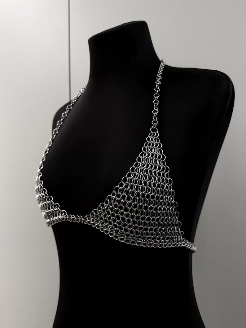 May include: A silver chainmail bikini top displayed on a black mannequin. The top features a halter neck design and triangular cups, constructed entirely of small, interlocking metal rings. The metallic garment is shown against a neutral background.