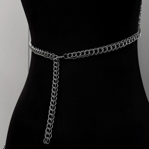 May include: A silver chain belt with a series of interlocking circular links. The belt is draped on a black mannequin, showcasing its design. A longer chain hangs down from the center of the belt, adding a decorative element. The belt is a fashion accessory.