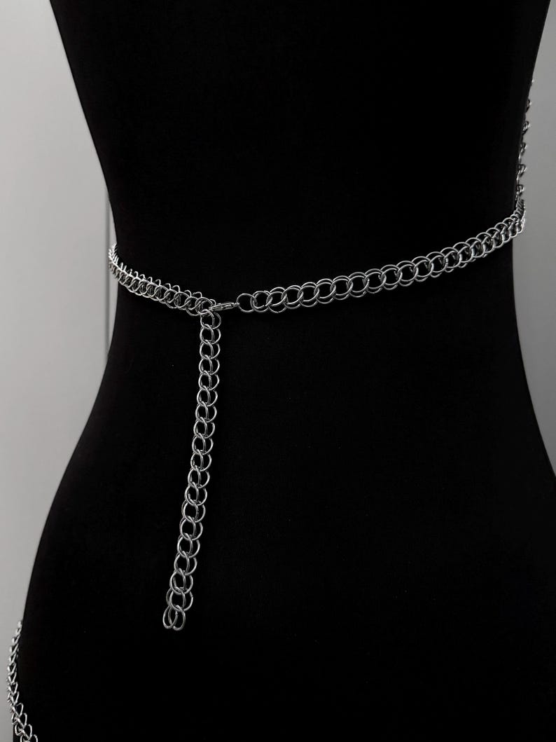 May include: A silver chain belt with a series of interlocking circular links. The belt is draped on a black mannequin, showcasing its design. A longer chain hangs down from the center of the belt, adding a decorative element. The belt is a fashion accessory.