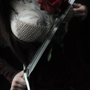 May include: A close-up shot of a person holding a long, silver sword and a red rose. The person is wearing a chainmail top and has intricate tattoos on their chest and arm. The background is dark.