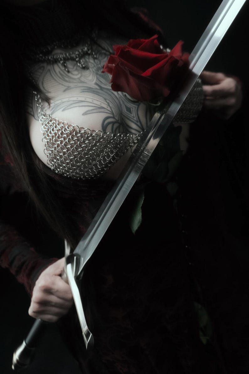 May include: A close-up shot of a person holding a long, silver sword and a red rose. The person is wearing a chainmail top and has intricate tattoos on their chest and arm. The background is dark.