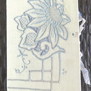 Può includere: Un diario rivestito in tessuto color crema con un intricato ricamo floreale azzurro. La copertina presenta un grande motivo floreale centrale con dettagli traforati, bordi geometrici e una piccola chiusura a bottone sul lato.