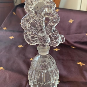 Puede incluir: Un frasco de perfume de vidrio transparente con un tapón de diseño floral. El frasco tiene un cuerpo acampanado con estrías.