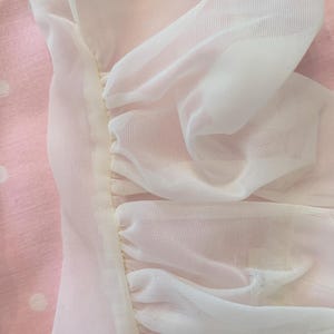 Peut inclure: Gros plan sur une section de tissu blanc transparent, froncé et cousu avec un fil jaune clair. Le tissu est superposé, créant un effet texturé. Le fond est rose clair avec des pois blancs.