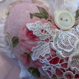 May include: A pink heart-shaped pillow with white lace trim and a white button. The pillow has a floral pattern with pink roses and green leaves.