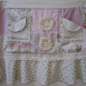Puede incluir: Un delantal de patchwork rosa y blanco con ribete de encaje y rosetas de tela. El delantal tiene un dobladillo con volantes y varios bolsillos.