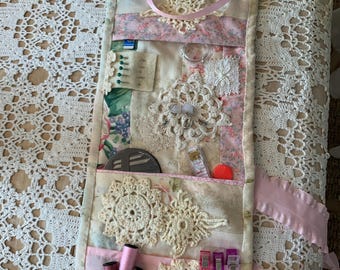 May include: A pink and white fabric roll-up sewing kit with various compartments. The kit includes scissors, needles, thread, bobbins, and other sewing supplies. It is decorated with lace and ribbons.