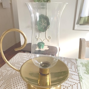 May include: A brass candle holder with a clear glass hurricane shade. The candle holder has a circular base and a curved handle. The glass shade is decorated with green floral accents. The candle holder sits on a white lace tablecloth.