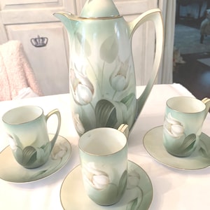 Puede incluir: Un juego de té de porcelana vintage con una cafetera alta con tapa y tres tazas a juego con platillos. El juego está pintado con un delicado diseño floral en tonos verdes y blancos, con ribetes dorados.