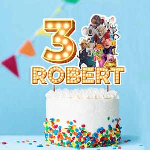 Puede incluir: Una tarta blanca glaseada con chispas de colores en la base. La tarta está coronada con un adorno decorativo con el número "3" y el nombre "ROBERT" en letras iluminadas, junto con personajes de dibujos animados.