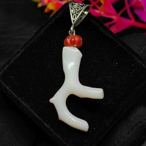 May include: A white coral pendant with a branch-like design, accented by a small red bead and a decorative silver bail. The pendant is displayed on a black surface, showcasing its natural form and color.