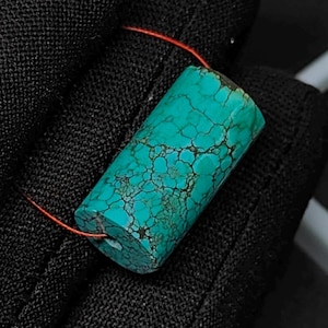 Puede incluir: Una cuenta cilíndrica de turquesa que presenta un color verde azulado vibrante con un patrón de matriz de telaraña de color marrón oscuro en toda la piedra. La cuenta está ensartada en un cordón rojo fino, mostrando su textura natural.
