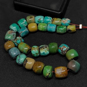 Pode incluir: Um fio de contas de turquesa irregulares em forma de barril em vários tons de azul, verde e castanho. As contas apresentam padrões de matriz naturais e estão enfiadas num fio vermelho fino, exibindo uma mistura de texturas polidas e mate.