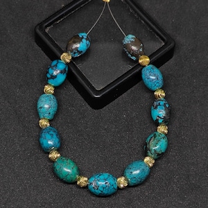 May include: A beaded necklace featuring oval-shaped turquoise stones with dark matrix patterns, interspersed with small, round, textured gold-tone metal spacer beads. The beads are strung on a thin, clear filament wire, creating a circular arrangement.