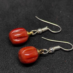 May include: A pair of red coral earrings with silver hooks. Each earring features a carved, oval-shaped coral bead with vertical grooves. The beads are attached to silver hooks with gold-colored metal accents. The earrings are displayed against a dark background.