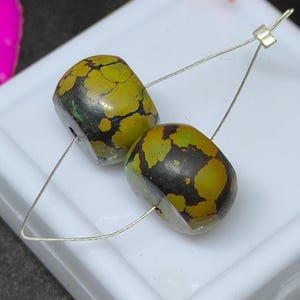 Könnte beinhalten: Zwei fassförmige Perlen mit einem gesprenkelten Muster aus Olivgrün und Schwarz. Die Perlen sind auf einem dünnen Silberdraht aufgereiht und vor einem weißen Hintergrund platziert. Die Perlen sind wahrscheinlich für die Schmuckherstellung.