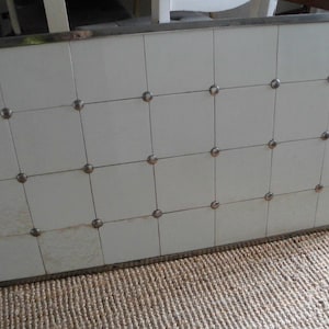 Könnte beinhalten: Ein rechteckiger, weiß gefliester Tisch mit einem silbernen Metallrahmen und dekorativen silbernen Nieten. Der Tisch steht auf einem gewebten Jute-Teppich. Der Tisch ist etwa 91 cm lang und 61 cm breit.