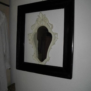 Könnte beinhalten: Ein dekorativer Wandspiegel. Er zeigt einen weißen, verzierten, oval geformten Spiegel, der in einem größeren schwarzen rechteckigen Rahmen eingefasst ist. Der Spiegel ist an einer weißen Wand montiert.