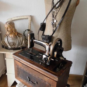 Könnte beinhalten: Eine antike Nähmaschine mit einem Holzschrank und verzierten Metalldetails. Die Maschine ist schwarz und goldfarben, mit einem großen Schwungrad. Eine Büste einer religiösen Figur und eine Schaufensterpuppe mit Accessoires befinden sich im Hintergrund.
