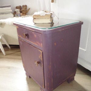 Könnte beinhalten: Ein Nachttisch im Vintage-Stil in Lila mit Glasplatte. Der Nachttisch hat eine Schublade und eine Tür mit einem Knopf. Die Farbe hat ein Used-Finish. Zwei Bücher liegen oben auf dem Nachttisch.