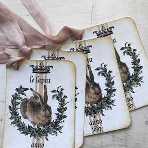 May include: Four vintage-style tags with a rabbit illustration, text, and a crown design. The tags are cream-colored with a distressed look, featuring a brown rabbit inside a green wreath. The text "Le lapin" and "anno 1867" are printed on the tags. A light brown ribbon is tied to the tags.
